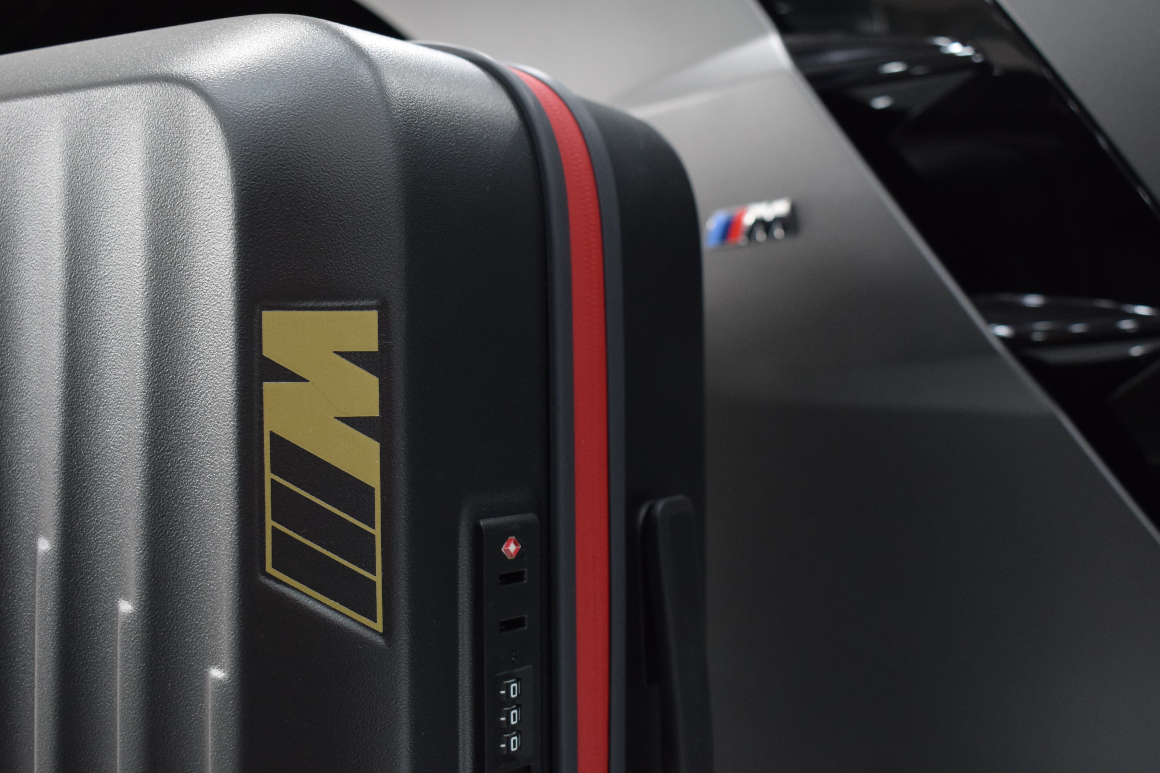 Valija rígida BMW M con franja roja junto a un BMW en el concesionario